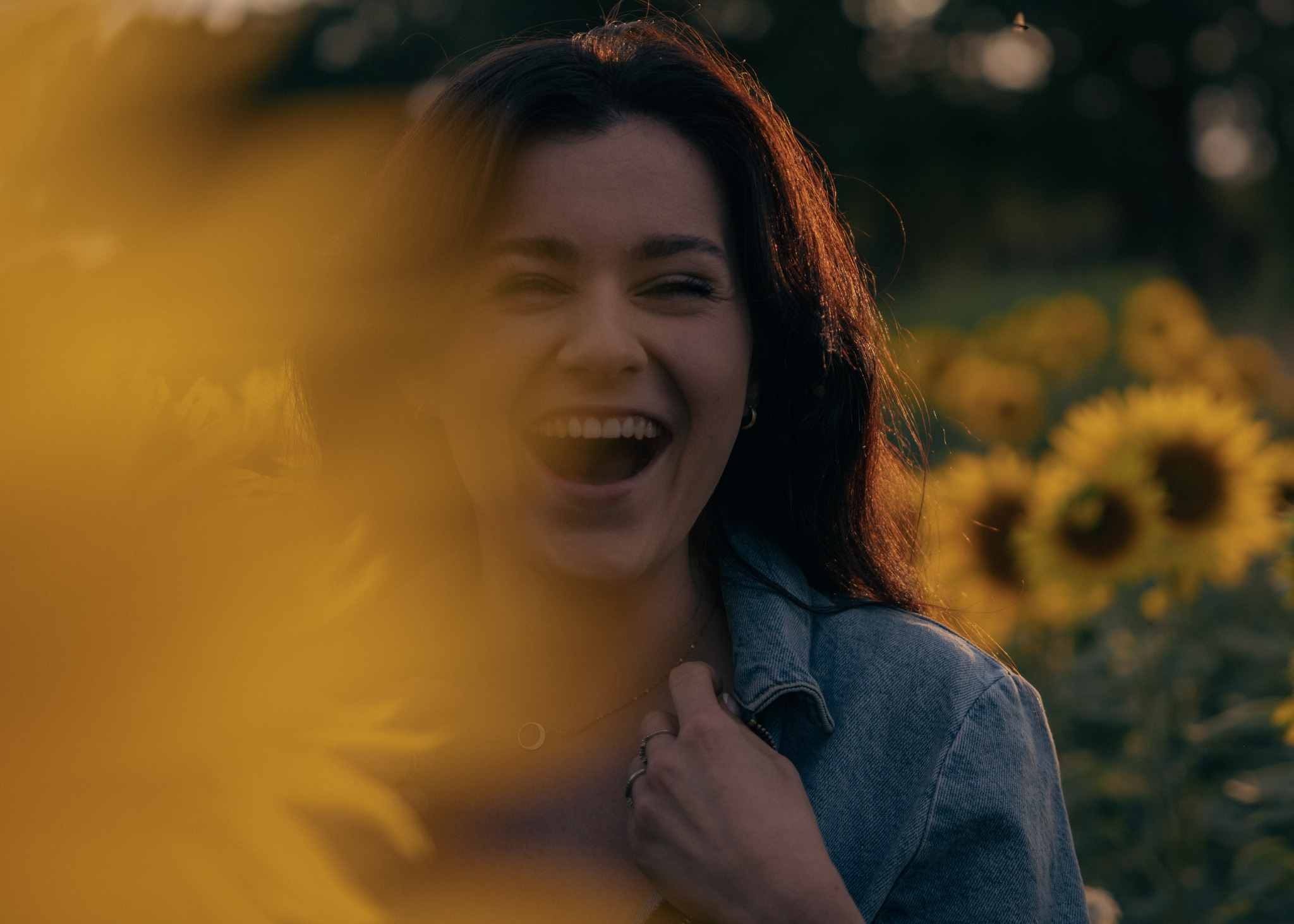 Rozesmiana brunetka w dżinsowej kurtce, częściowo zasłonięta przez kwiat słonecznika, na tle pola słoneczników.