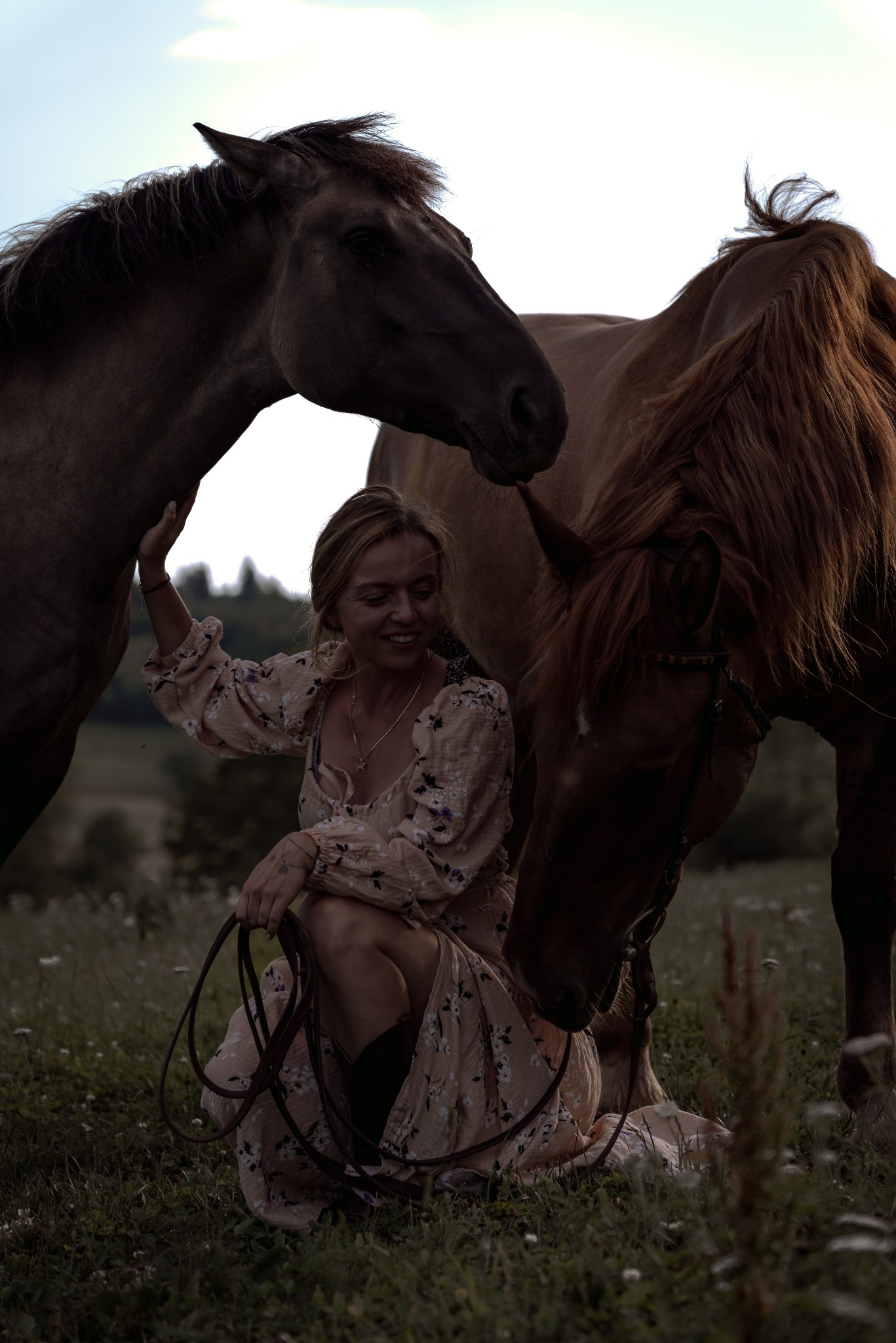 Uśmiechnięta kobieta w kwiecistej sukience kuca na łące, obejmując dwa konie o brązowej sierści; jeden z koni ma ciemniejszą grzywę.