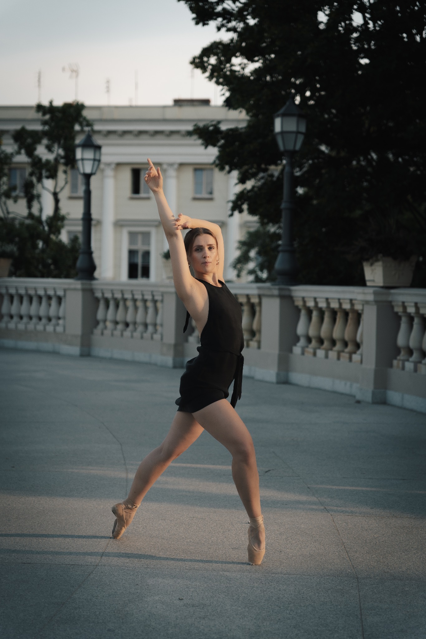 Baletnica w czarnym stroju pozuje na tle klasycystycznej architektury, unosząc rękę w górę podczas występu.