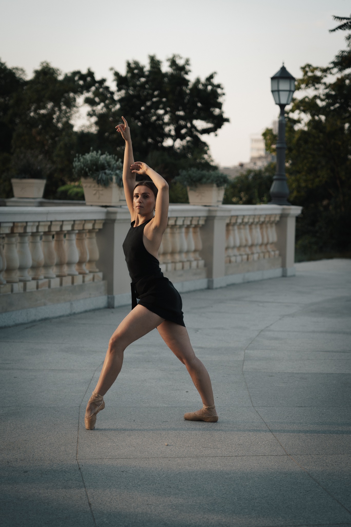 Kobieta w czarnej sukience baletowej pozuje na tle miejskiego parku z balustradami i latarnią, unosząc ręce w górę.