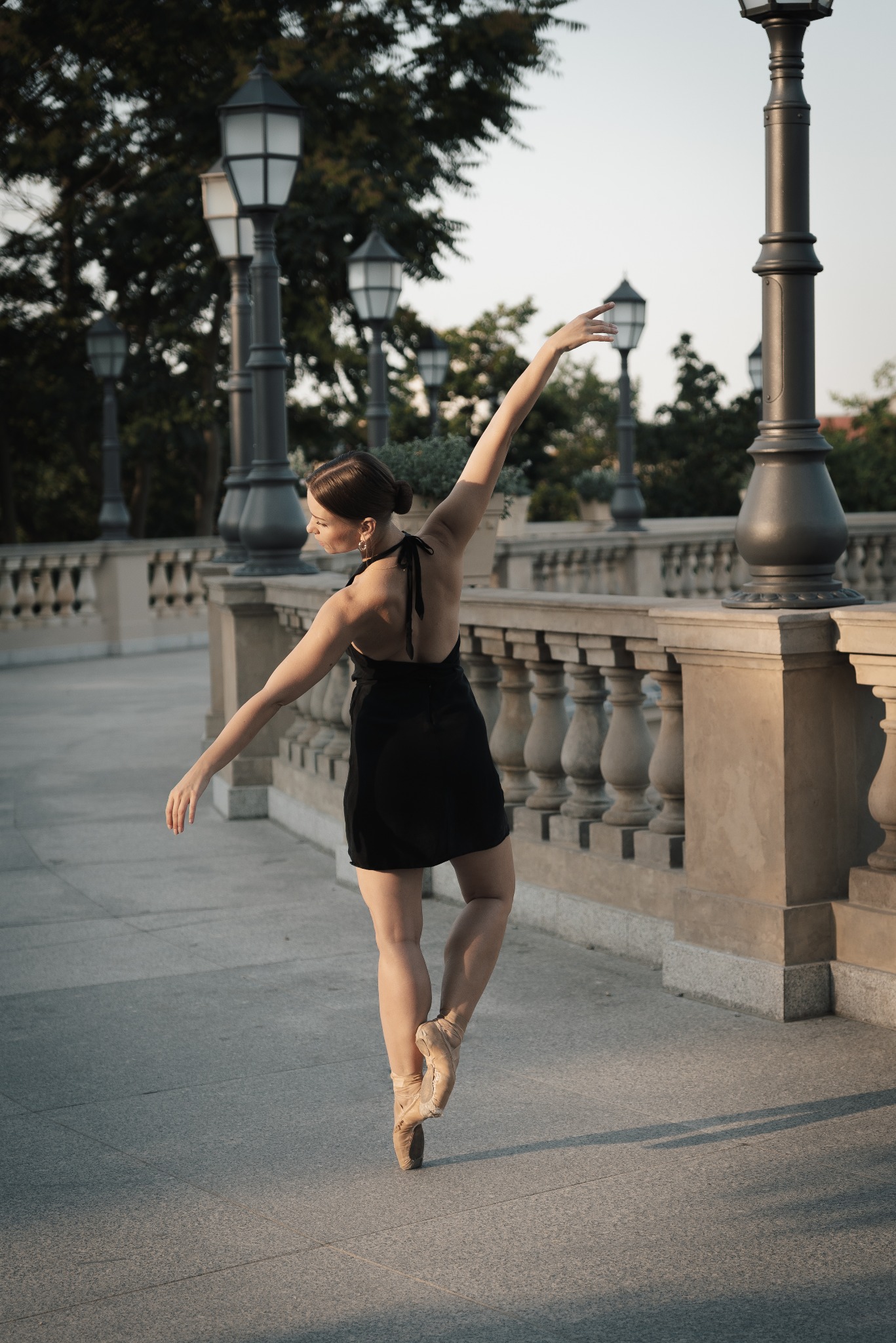 Baletnica w czarnej sukience pozuje na tle balustrady i latarni w parku, stojąc na pointach.