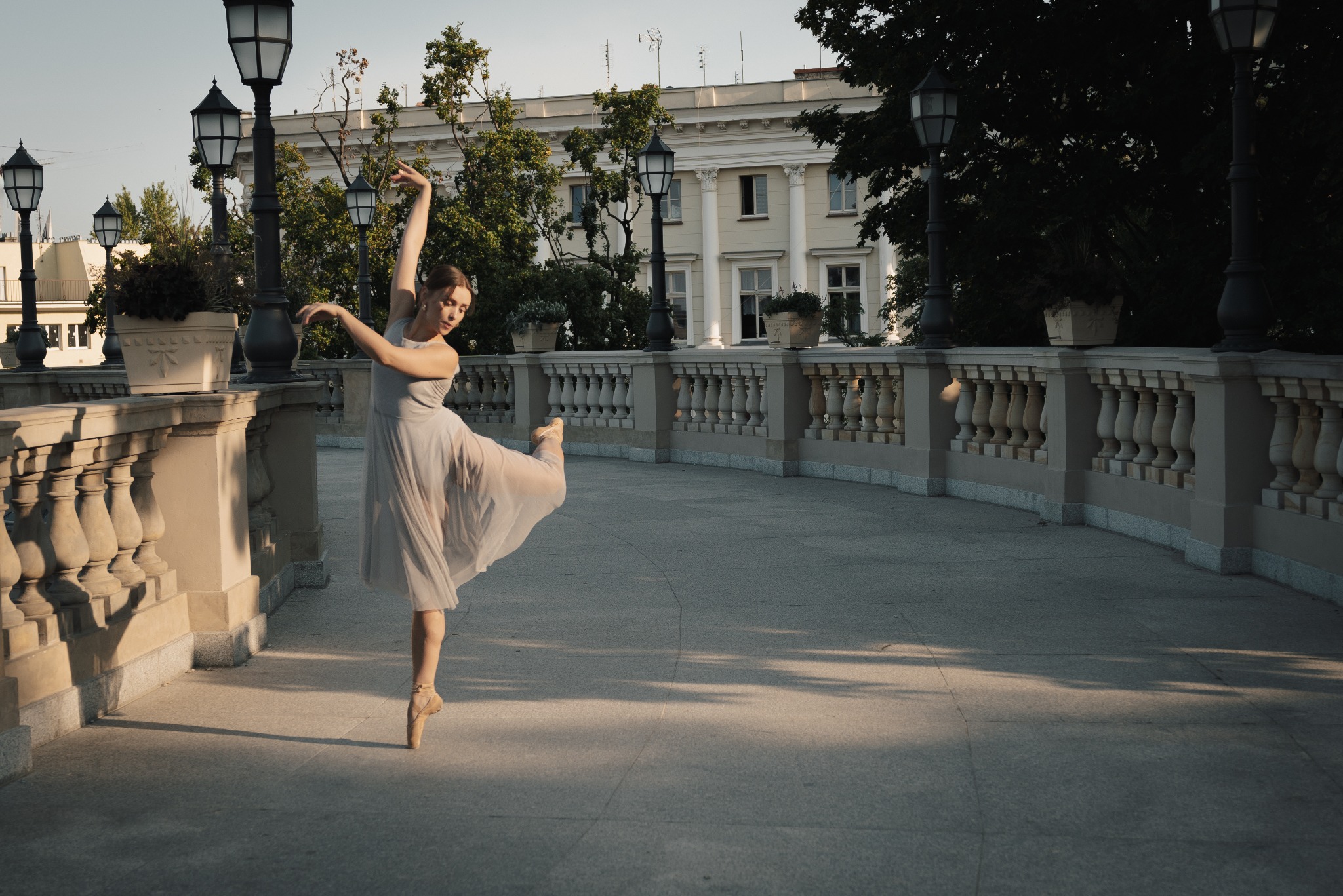 Baletnica w zwiewnej sukience wykonuje taniec na tle klasycystycznej architektury i latarni.