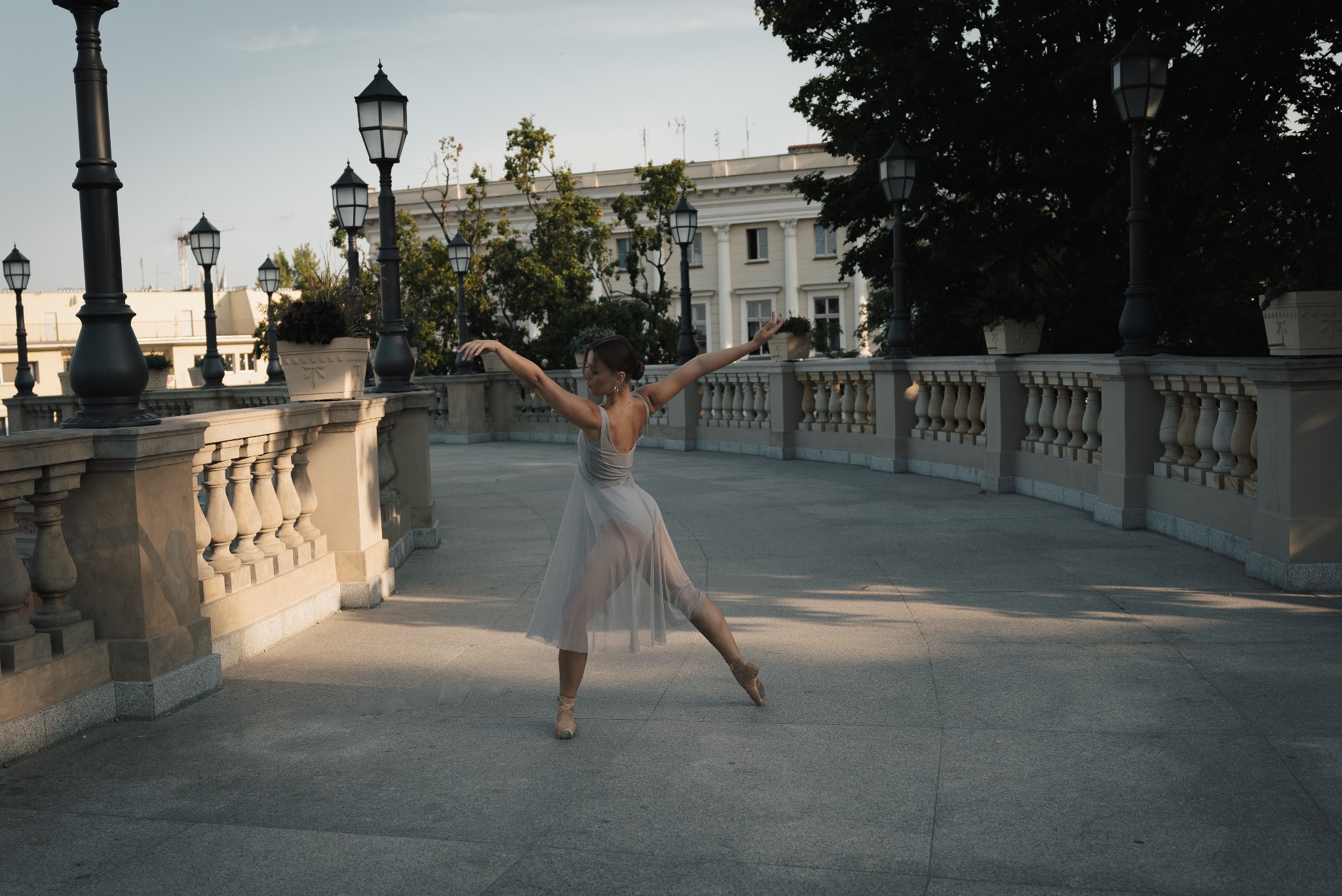 Baletnica w zwiewnej sukience wykonuje taniec na kamiennym moście z ozdobnymi latarniami i balustradami, w tle jasny budynek.