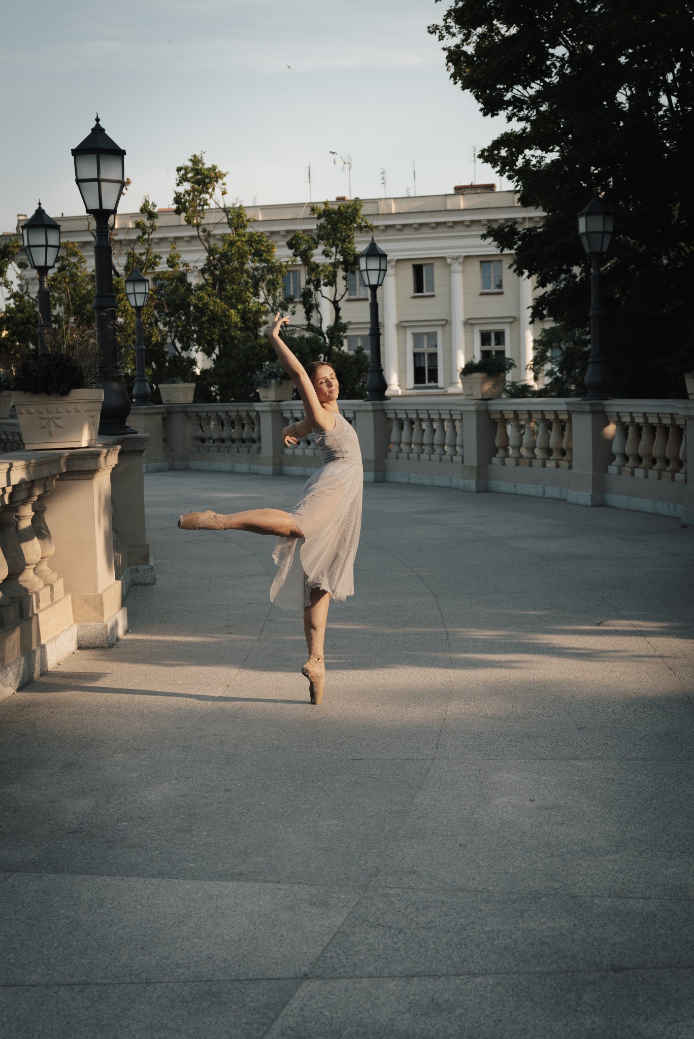 Baletnica w zwiewnej sukience wykonuje figurę na tle balustrady i latarni w miejskim parku, z budynkiem w tle.
