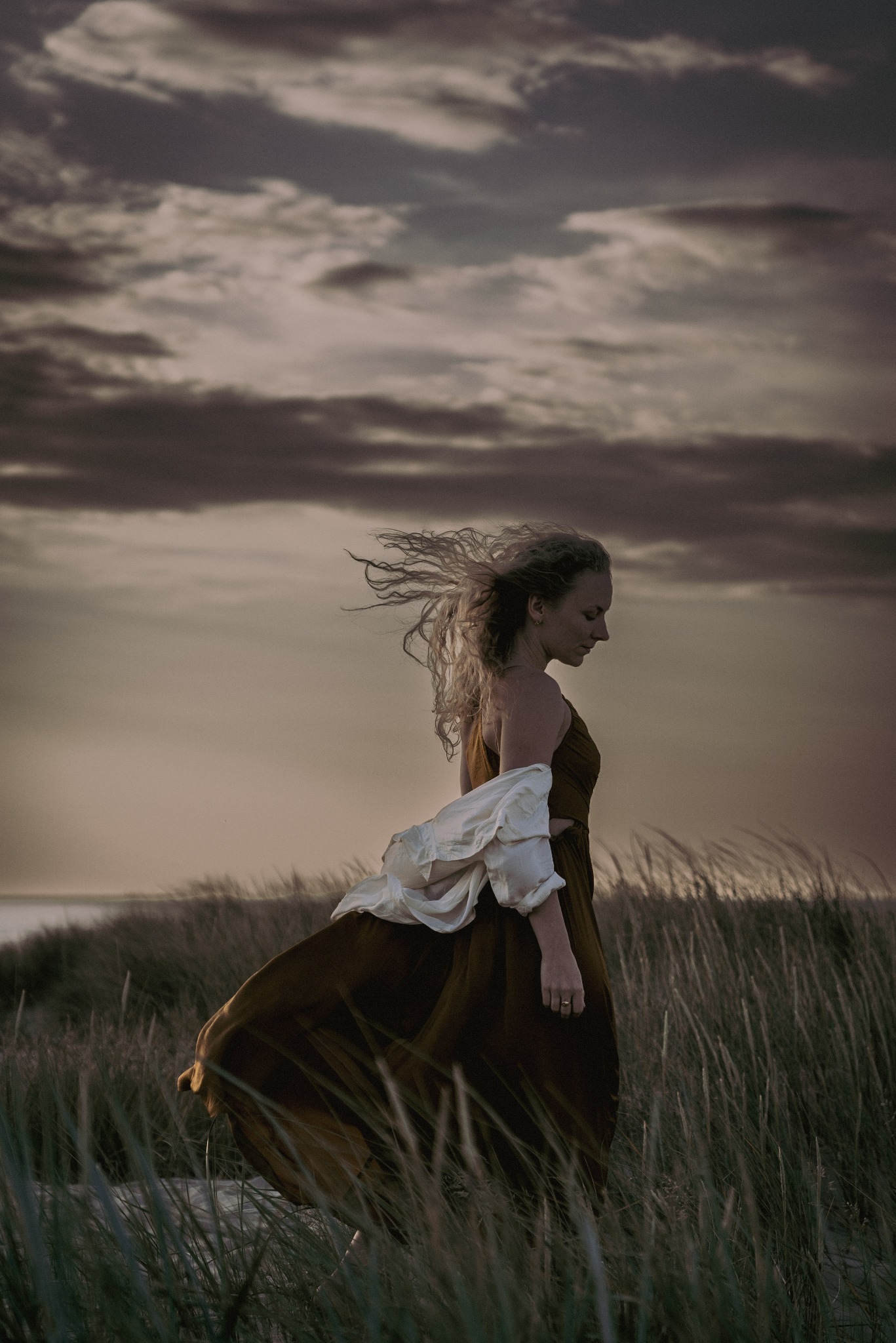 Kobieta w zwiewnej sukience i białej bluzce idzie przez trawę na tle zachmurzonego nieba i morza w oddali.