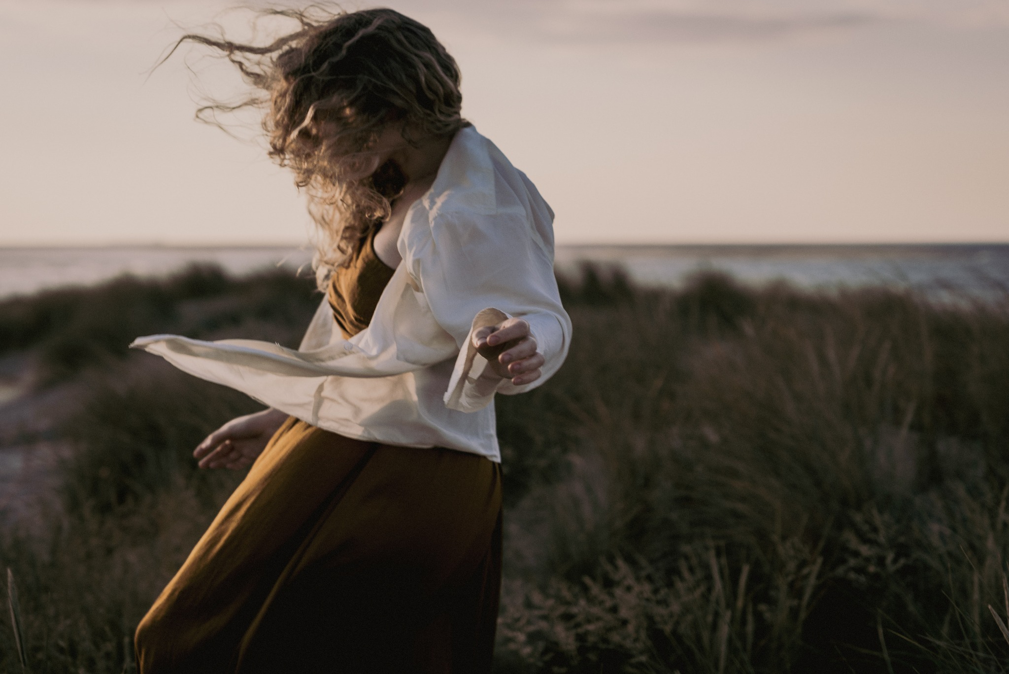 Kobieta w zwiewnej sukience i białej koszuli tańczy na plaży, włosy rozwiane przez wiatr, w tle morze i piasek o zmierzchu.
