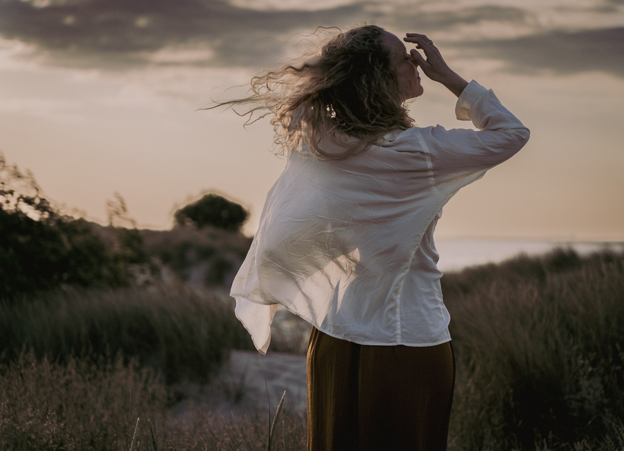 Kobieta w białej koszuli i długiej spódnicy, stojąca tyłem na tle zachodzącego słońca, z włosami rozwianymi na wietrze, dotyka czoła