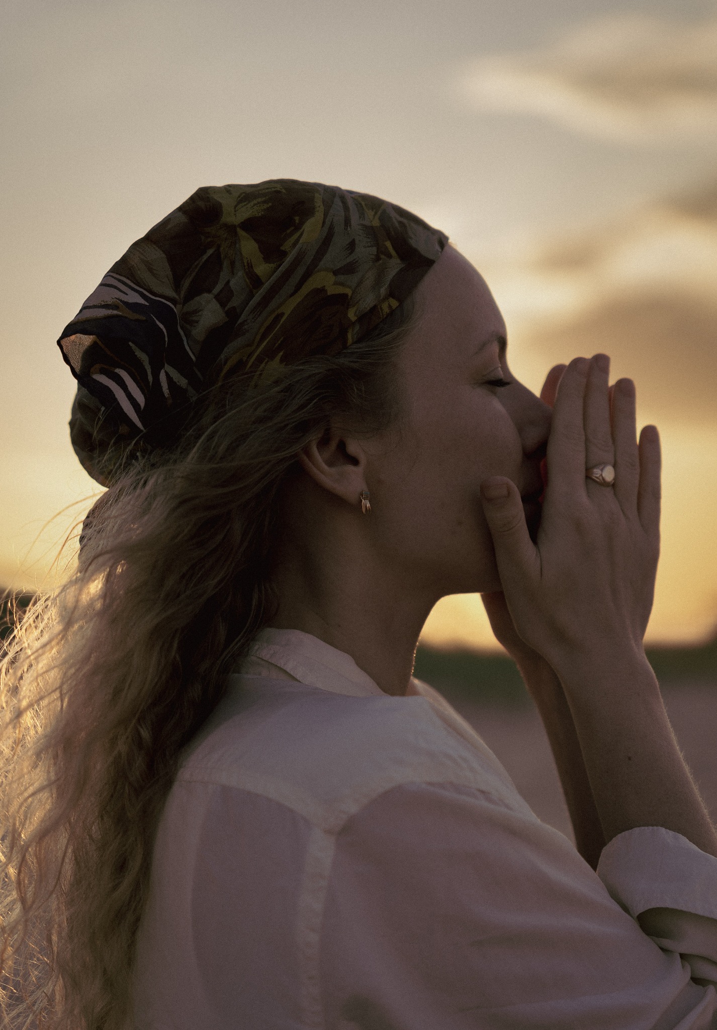 Portret kobiety z zamkniętymi oczami, dłońmi przy twarzy i chustą na głowie, w ciepłym świetle zachodzącego słońca.