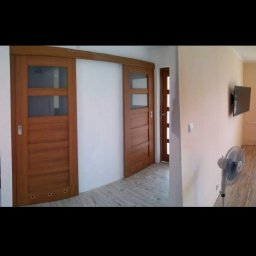 Evry-Bud - Panoramiczny widok wnętrza po remoncie: drewniane drzwi przesuwne i standardowe, nowa podłoga, świeżo pomalowane ściany w odcieniach beżu, wiszący telewizor, wentylator stojący i widok na kuchnię...