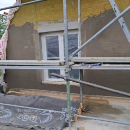 Evry-Bud - Fragment elewacji budynku w trakcie prac: ocieplenie z widoczną wełną mineralną, częściowo pokryte szarą warstwą tynku, okno zabezpieczone folią, rusztowanie z niebieskimi rękawicami roboczymi,...