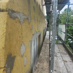 Evry-Bud - Elewacja budynku w trakcie ocieplania, widoczna żółta wełna mineralna z fragmentami siatki i zaprawy, rusztowanie wzdłuż ściany, okna zabezpieczone folią.