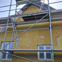 Evry-Bud - Dom w trakcie ocieplania z widocznymi rusztowaniami, oknami zabezpieczonymi folią i częściowo nałożoną warstwą izolacyjną z wełny mineralnej, zabezpieczoną folią Rockwool, z widocznymi fragmentami...