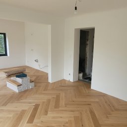 Evry-Bud - Jasne, nowe panele ułożone w jodełkę w przestronnym, odświeżonym pokoju z białymi ścianami. W tle widoczne okno z zielenią na zewnątrz i otwarte przejście do kolejnego pomieszczenia w trakcie prac...