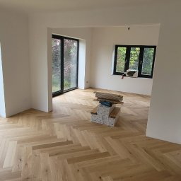 Evry-Bud - Jasny, drewniany parkiet ułożony we wzór jodełki w nowym, przestronnym pokoju z dużymi oknami i stosem paneli podłogowych.