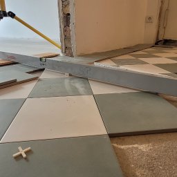 Evry-Bud - Układanie szachownicy z szaro-białych płytek podłogowych, poziomnica Bauhaus z napisem 'S.P.', krzyżyki dystansowe, widoczny fragment statywu laserowego i nierówna powierzchnia podłoża.