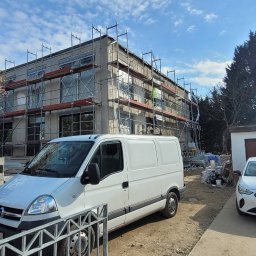 Evry-Bud - Dwupiętrowy budynek w trakcie prac elewacyjnych, otoczony rusztowaniami i zabezpieczony folią, z zaparkowanym białym vanem i samochodem osobowym na posesji.