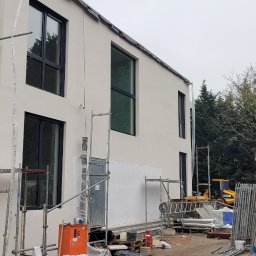 Evry-Bud - Nowoczesny dom w trakcie prac elewacyjnych: widoczne rusztowania, zabezpieczone okna folią, świeżo położony jasny tynk, teren budowy z narzędziami i materiałami, koparka w tle.