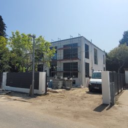 Evry-Bud - Nowoczesny dom jednorodzinny w trakcie prac elewacyjnych, widoczne rusztowania, palety z materiałami budowlanymi, teren wokół domu częściowo utwardzony, ogrodzenie panelowe.