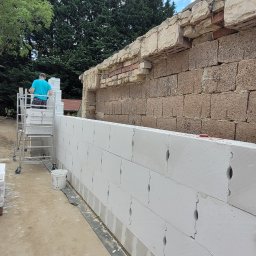 Evry-Bud - Budowa ściany z białych bloczków silikatowych, widoczny fragment starego muru z cegły i kamienia, pracownik na rusztowaniu oraz betoniarka i materiały izolacyjne w tle.