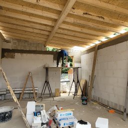 Evry-Bud - Wnętrze w trakcie prac wykończeniowych: pracownik na rusztowaniu nakłada zaprawę na ścianę z bloczków, widoczne nieotynkowane ściany, drewniana konstrukcja dachu oraz materiały budowlane...