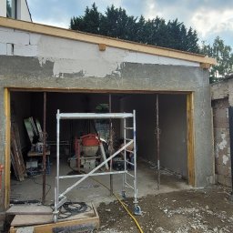 Evry-Bud - Budowa garażu: ściany w trakcie tynkowania, widoczne rusztowanie, mieszalnik i narzędzia wewnątrz, fragment posesji z numerem 49 w tle.