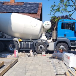 Evry-Bud - Niebieski betonomieszarka MAN na budowie, w tle dom z panelami słonecznymi i świeżo wylane betonowe schody wejściowe, widoczne narzędzia i materiały budowlane.