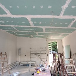 Evry-Bud - Wnętrze w trakcie prac wykończeniowych: sufit z płyt gipsowo-kartonowych z widocznymi łączeniami, wstępnie wyszpachlowane ściany, drabiny, narzędzia i materiały budowlane w pomieszczeniu z otwartym...