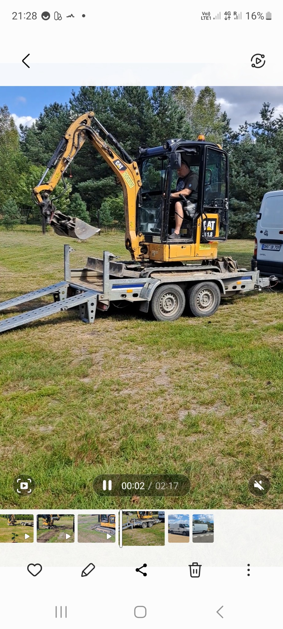 Żółta mini koparka CAT 1.8 na przyczepie z najazdami, operator siedzi w kabinie, w tle trawa i drzewa, obok biały bus.