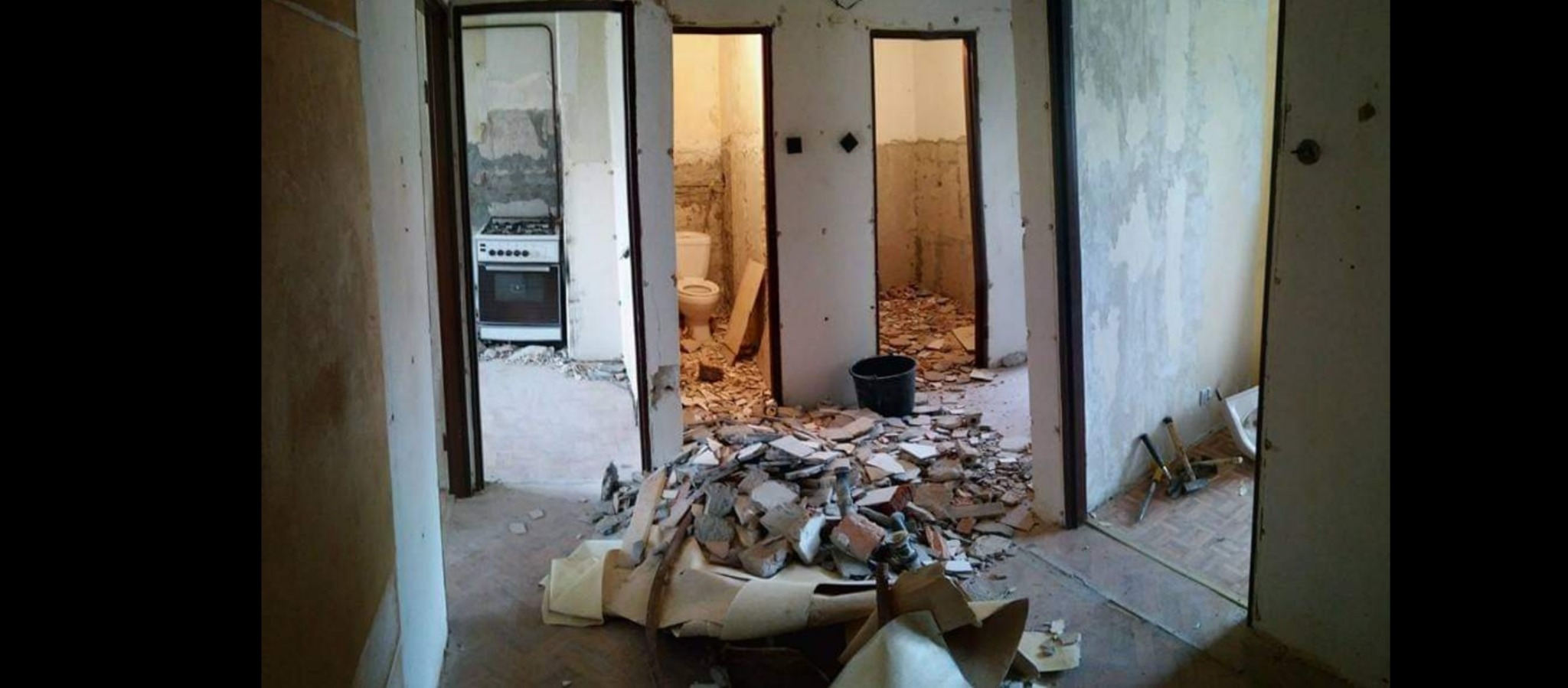 Wnętrze mieszkania w trakcie generalnego remontu: widoczne trzy otwarte przejścia do kuchni, łazienki i pokoju, gruz i resztki tapet na podłodze, ściany częściowo obdarte z okładzin, stara kuchenka...