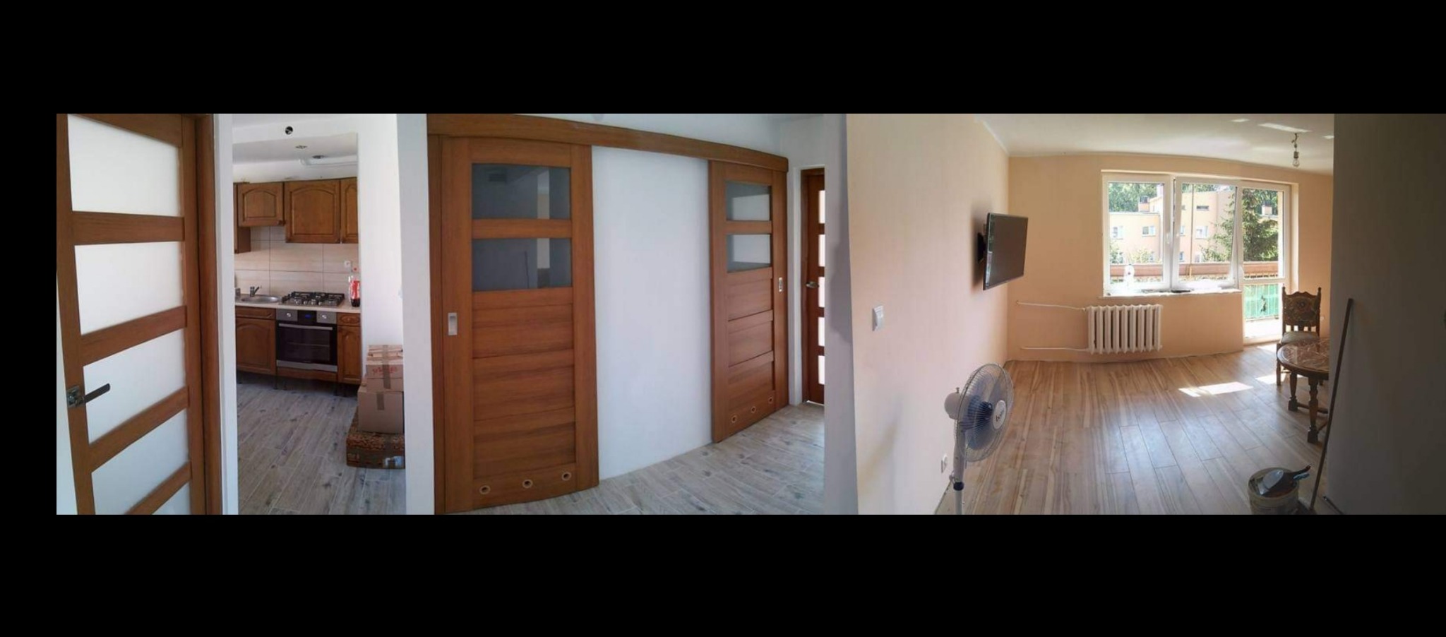 Panoramiczny widok wnętrza po remoncie: drewniane drzwi przesuwne i standardowe, nowa podłoga, świeżo pomalowane ściany w odcieniach beżu, wiszący telewizor, wentylator stojący i widok na kuchnię...