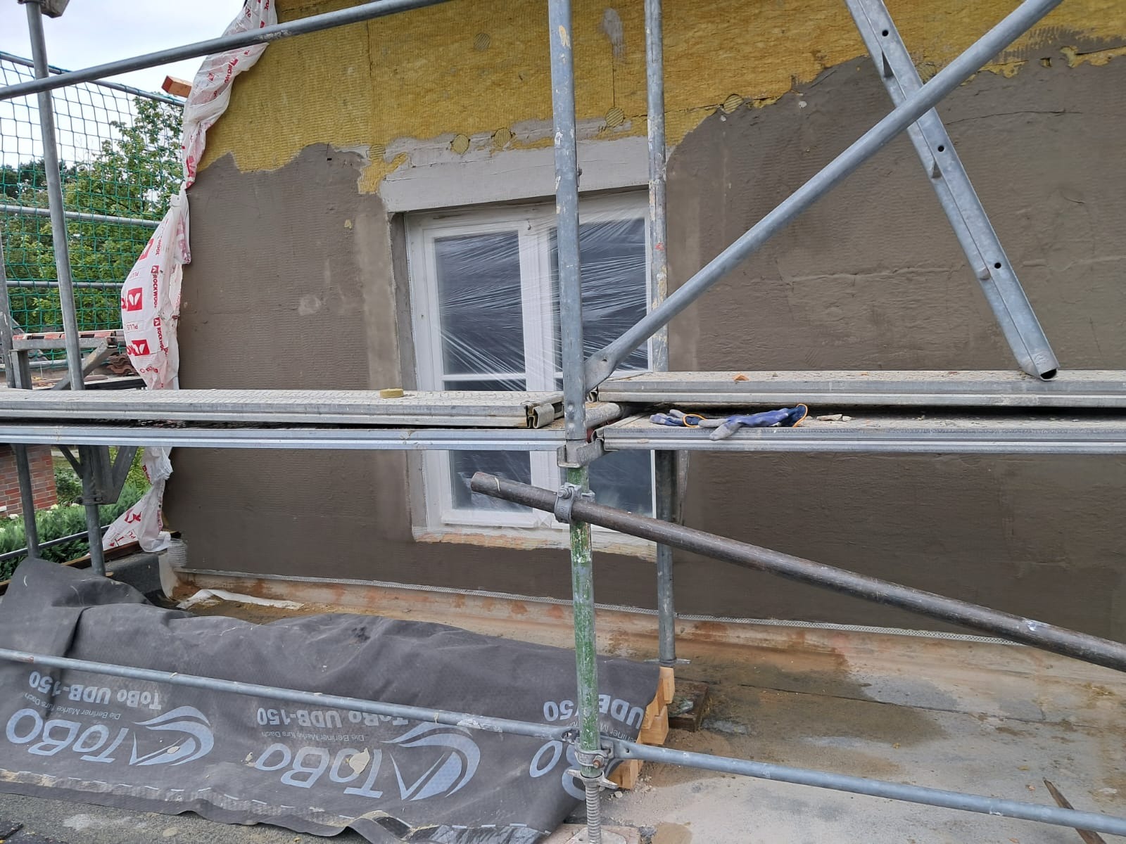 Fragment elewacji budynku w trakcie prac: ocieplenie z widoczną wełną mineralną, częściowo pokryte szarą warstwą tynku, okno zabezpieczone folią, rusztowanie z niebieskimi rękawicami roboczymi,...
