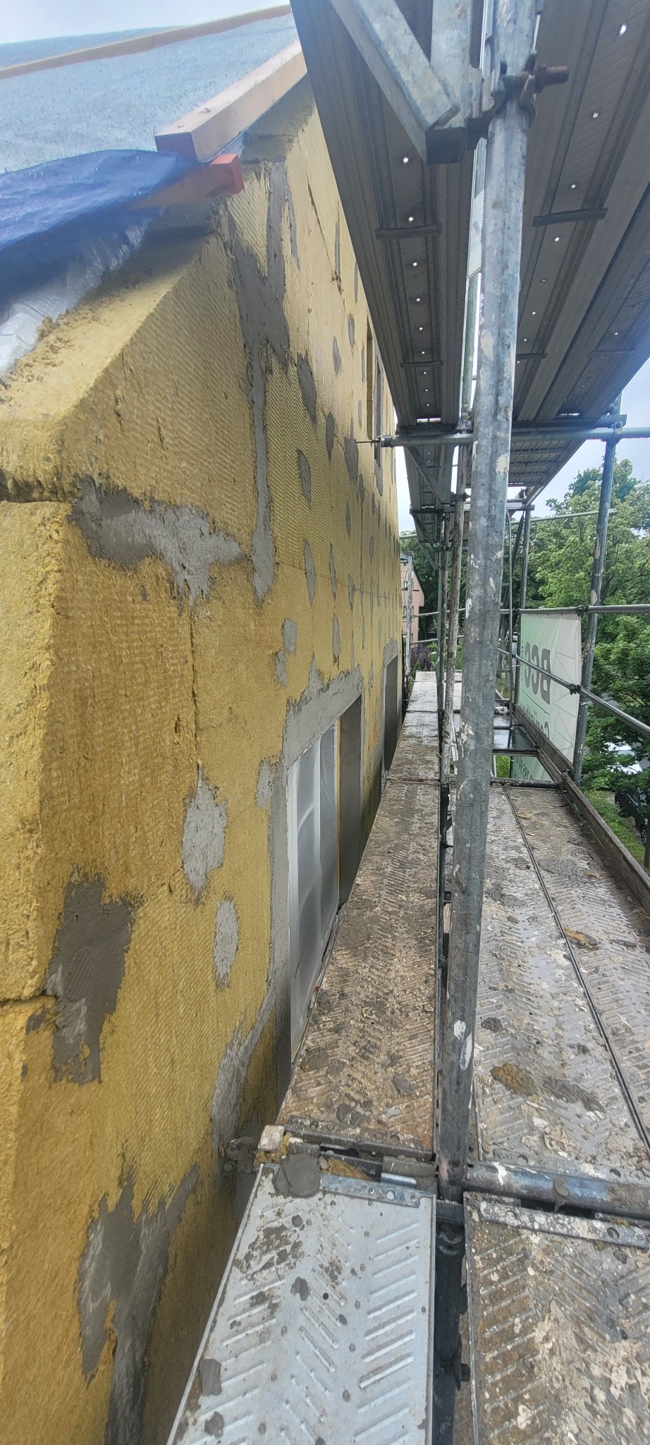 Elewacja budynku w trakcie ocieplania, widoczna żółta wełna mineralna z fragmentami siatki i zaprawy, rusztowanie wzdłuż ściany, okna zabezpieczone folią.