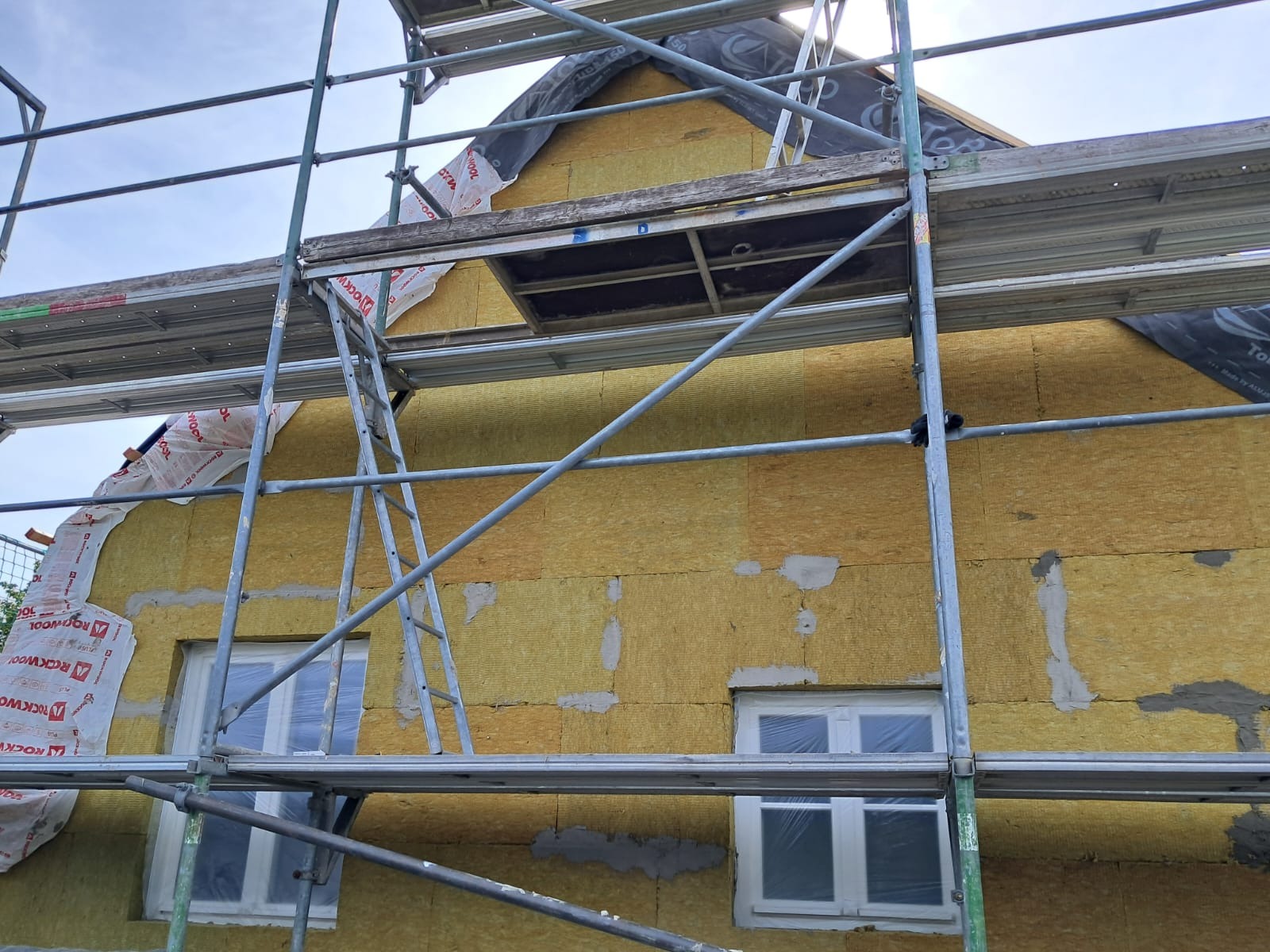 Dom w trakcie ocieplania z widocznymi rusztowaniami, oknami zabezpieczonymi folią i częściowo nałożoną warstwą izolacyjną z wełny mineralnej, zabezpieczoną folią Rockwool, z widocznymi fragmentami...