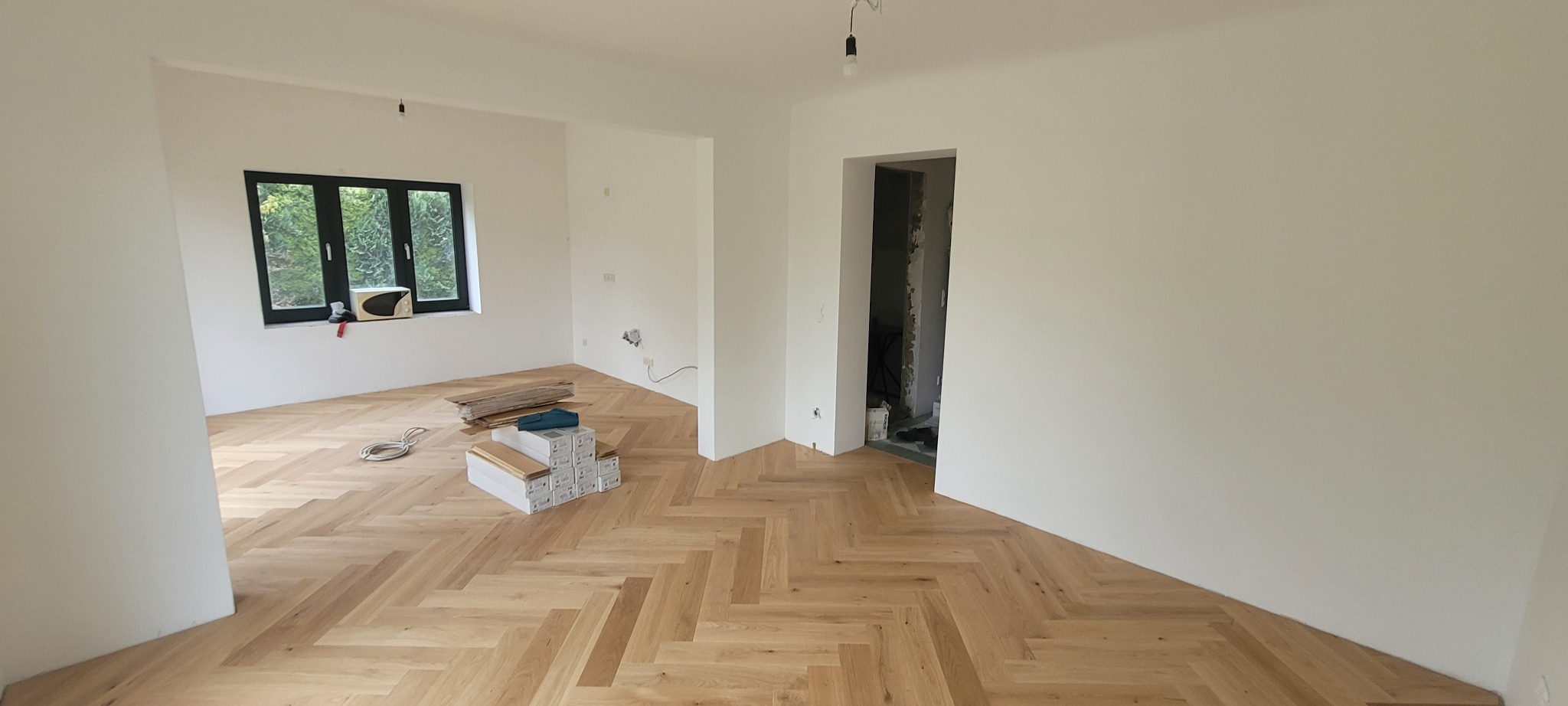 Jasne, nowe panele ułożone w jodełkę w przestronnym, odświeżonym pokoju z białymi ścianami. W tle widoczne okno z zielenią na zewnątrz i otwarte przejście do kolejnego pomieszczenia w trakcie prac...