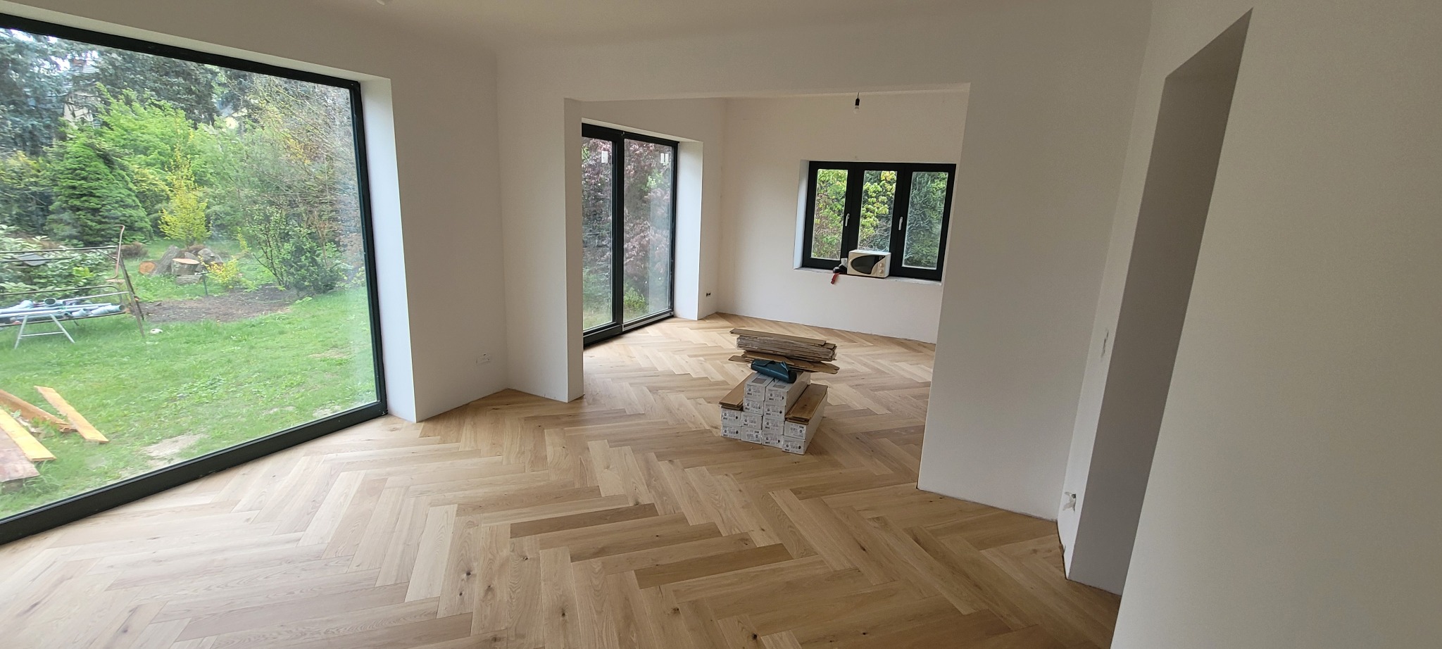 Jasny, drewniany parkiet ułożony we wzór jodełki w nowym, przestronnym pokoju z dużymi oknami i stosem paneli podłogowych.