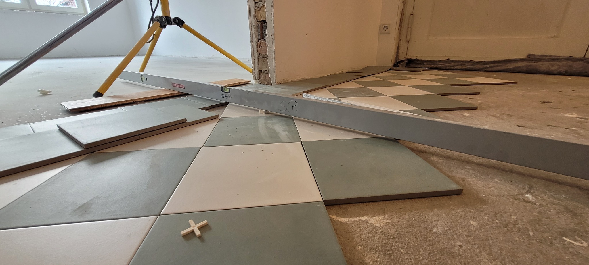 Układanie szachownicy z szaro-białych płytek podłogowych, poziomnica Bauhaus z napisem 'S.P.', krzyżyki dystansowe, widoczny fragment statywu laserowego i nierówna powierzchnia podłoża.