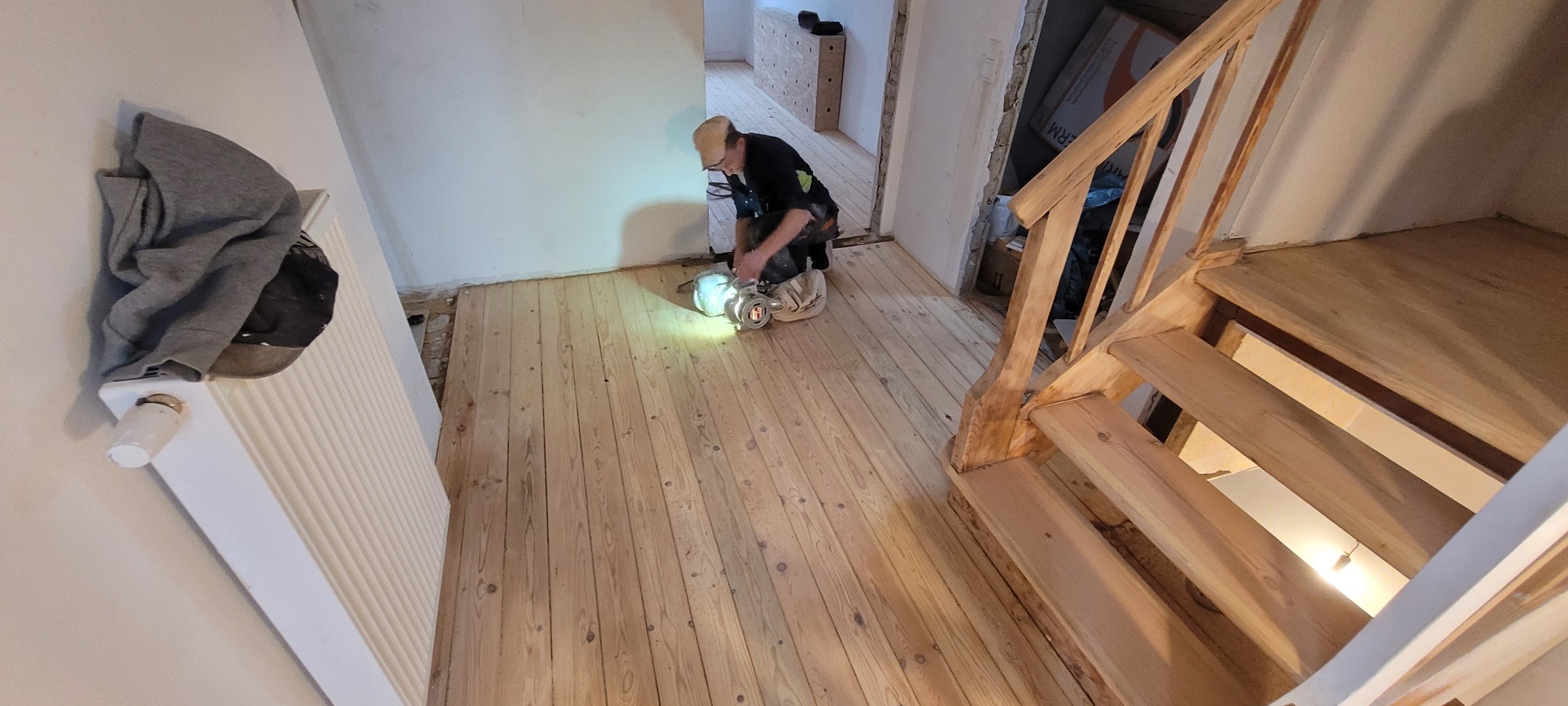 Renowacja drewnianej podłogi w trakcie: szlifierka w użyciu, widoczne schody i grzejnik z ubraniami.