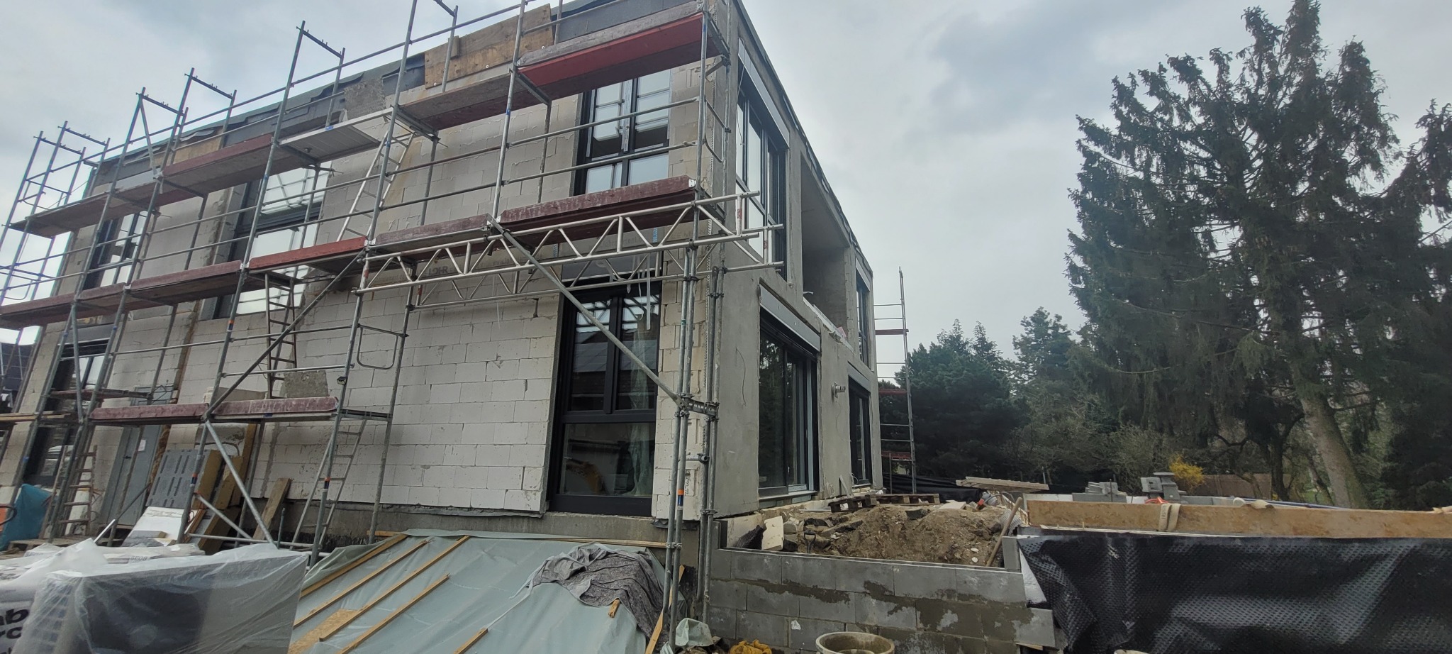 Budynek w trakcie budowy z widocznymi rusztowaniami, ścianami z białych bloczków i zamontowanymi oknami z czarnymi ramami, na tle drzew i pochmurnego nieba.