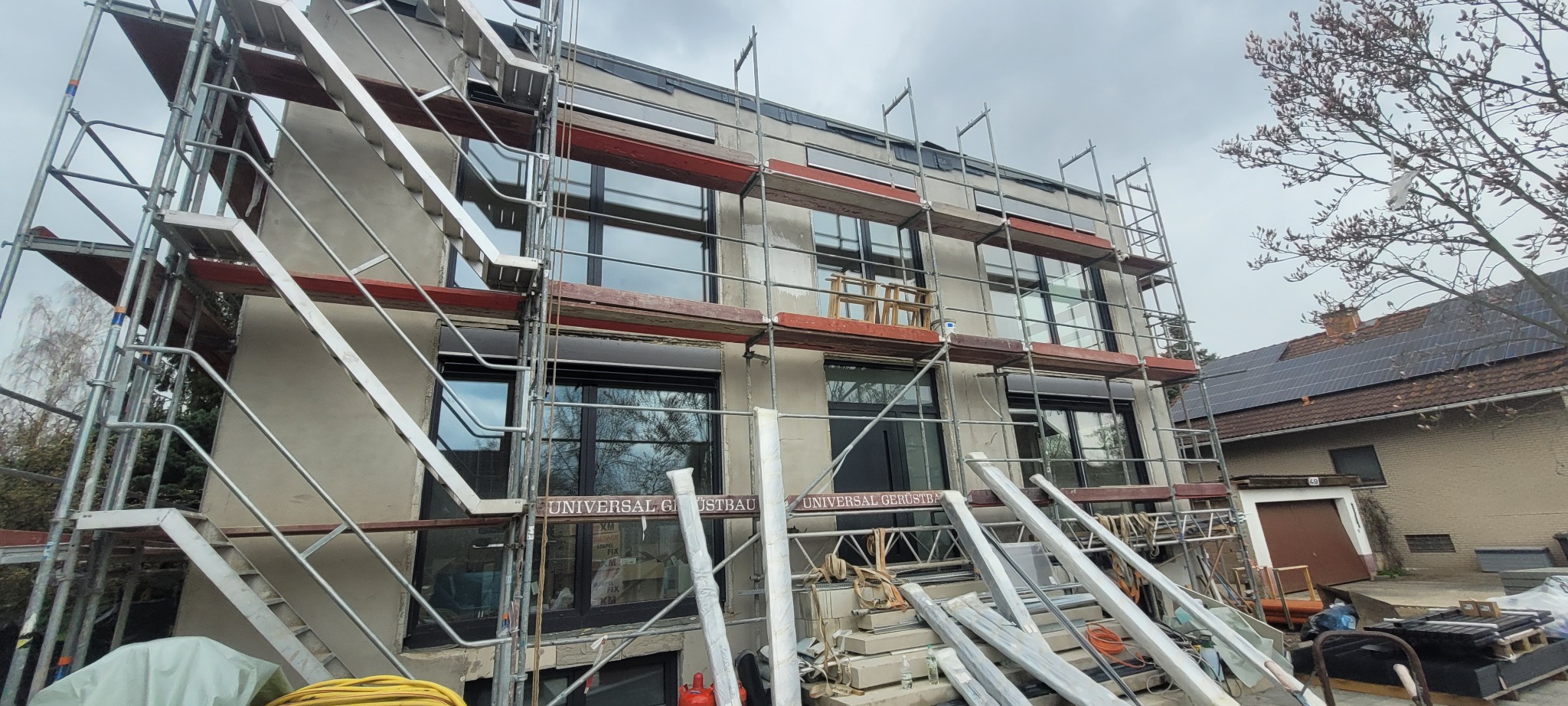 Budynek mieszkalny w trakcie prac elewacyjnych, widoczne rusztowania, okna z roletami zewnętrznymi, ściany w surowym tynku, przygotowane do dalszych prac wykończeniowych, w tle dom z panelami...