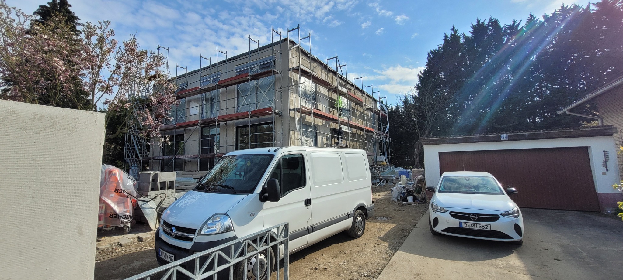 Dwupiętrowy budynek w trakcie prac elewacyjnych, otoczony rusztowaniami i zabezpieczony folią, z zaparkowanym białym vanem i samochodem osobowym na posesji.