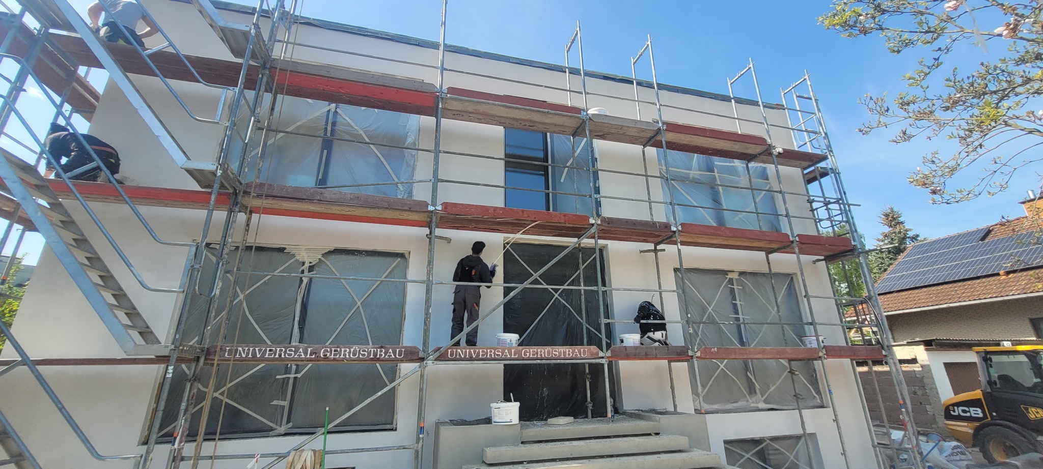 Budynek w trakcie prac elewacyjnych, osłonięte okna folią, rusztowania, pracownik w kombinezonie, widoczne wiadra z farbą, częściowo nałożona biała warstwa, w tle dom z panelami słonecznymi.