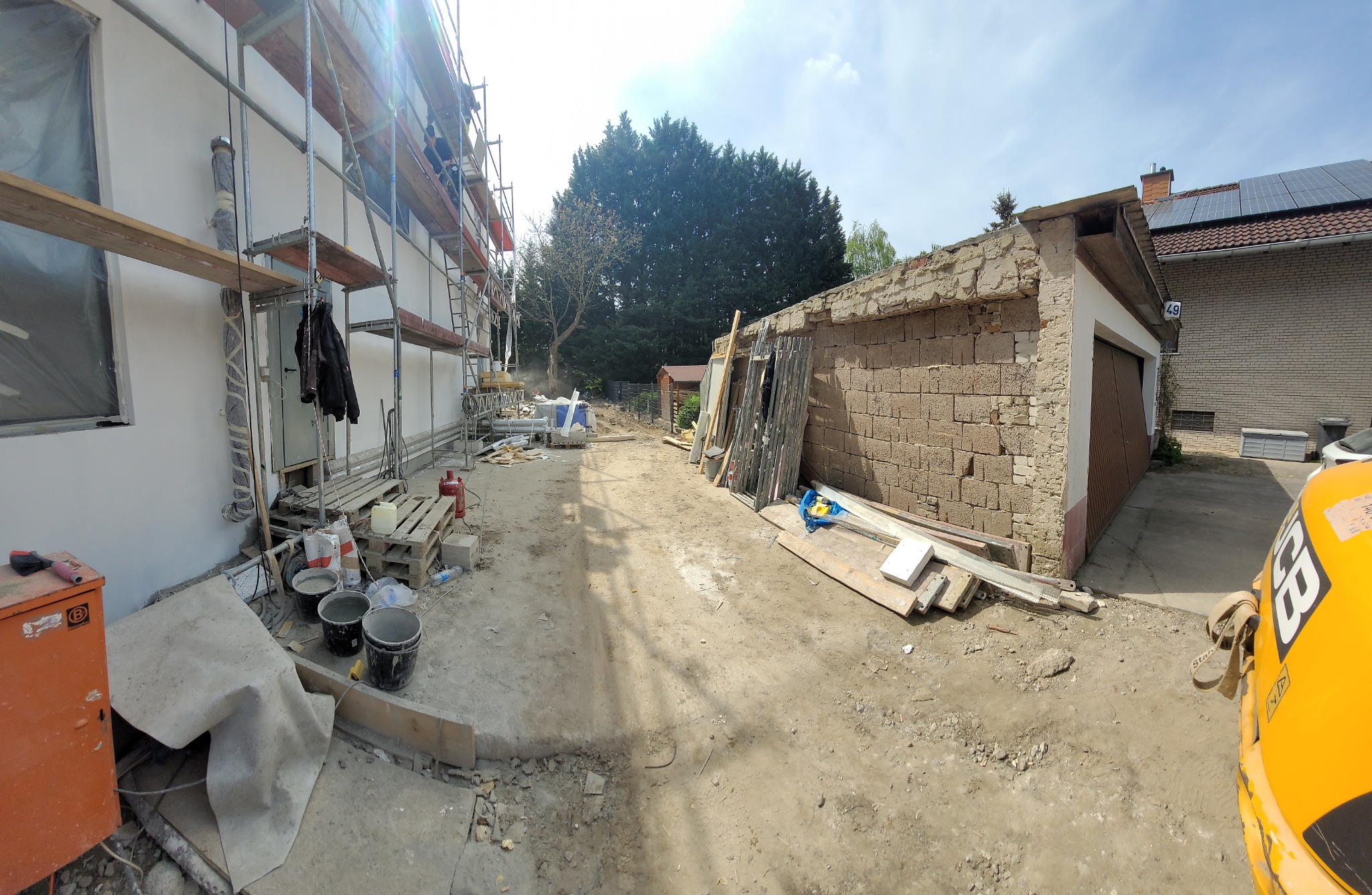 Ocieplanie elewacji budynku jednorodzinnego w trakcie realizacji, widoczne rusztowania, materiały budowlane, narzędzia i fragment muru oporowego. W tle drzewa i budynek z panelami słonecznymi.