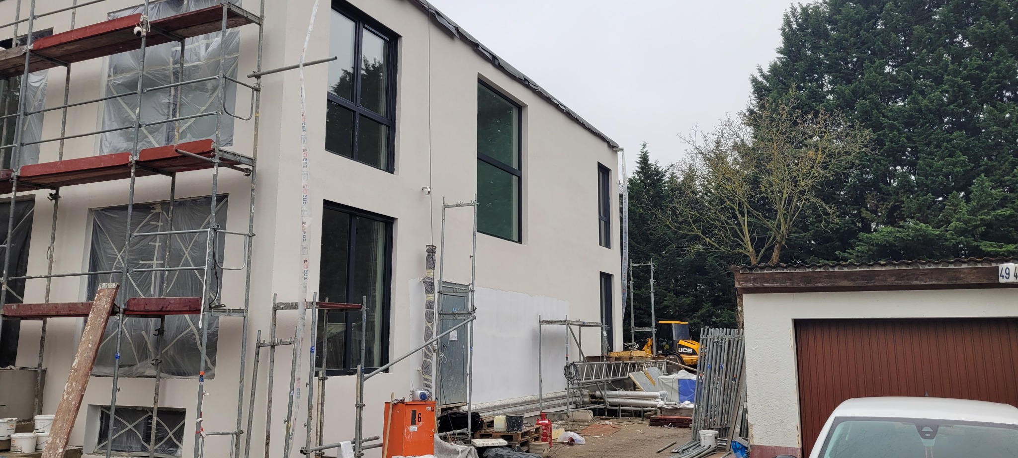 Nowoczesny dom w trakcie prac elewacyjnych: widoczne rusztowania, zabezpieczone okna folią, świeżo położony jasny tynk, teren budowy z narzędziami i materiałami, koparka w tle.