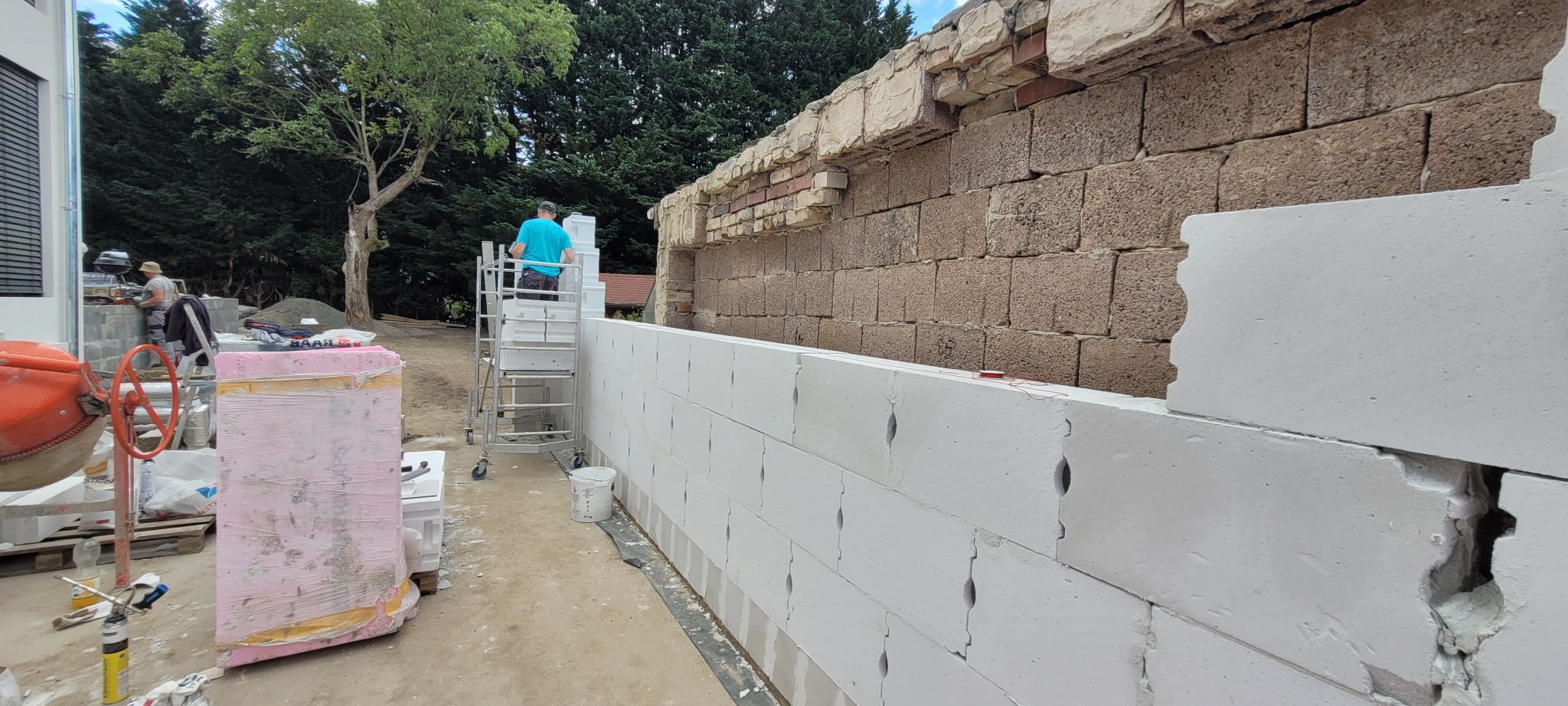 Budowa ściany z białych bloczków silikatowych, widoczny fragment starego muru z cegły i kamienia, pracownik na rusztowaniu oraz betoniarka i materiały izolacyjne w tle.