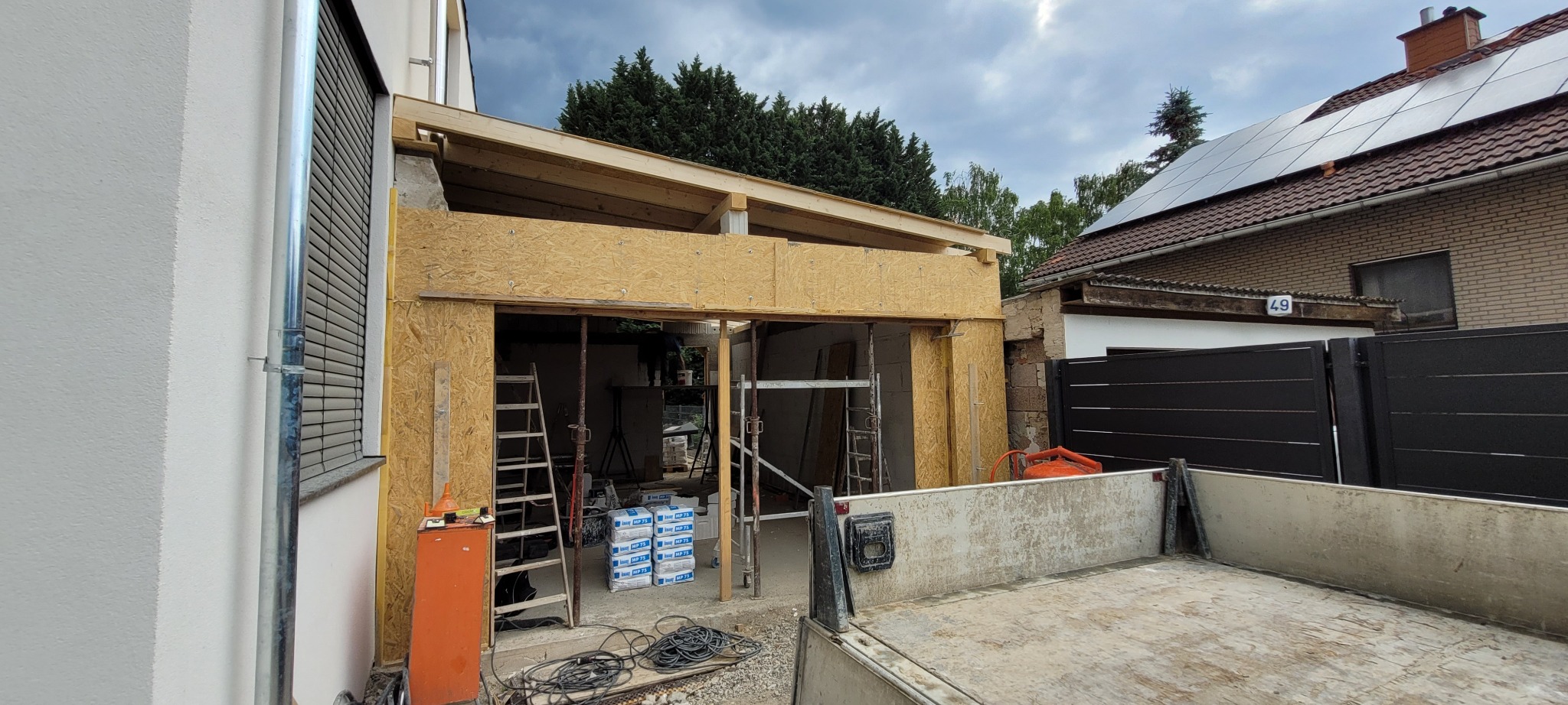 Budowa rozbudowy domu w trakcie: drewniane elementy konstrukcyjne, płyty OSB, rusztowania, drabina i zapas materiałów budowlanych widoczne na placu budowy.