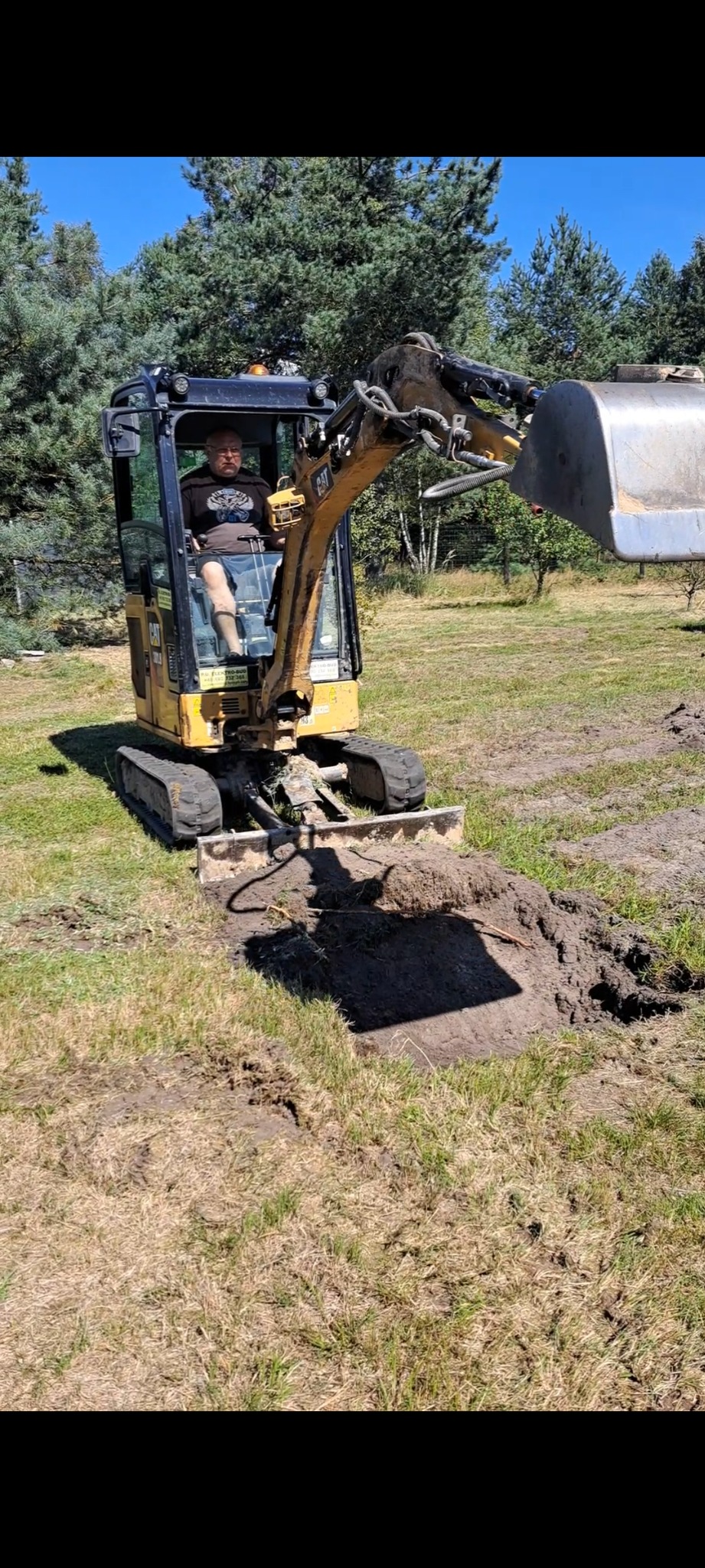 Operator w kabinie żółtej mini-koparki CAT, wykonujący wykop w trawiastym terenie pod budowę fundamentów lub instalacji.