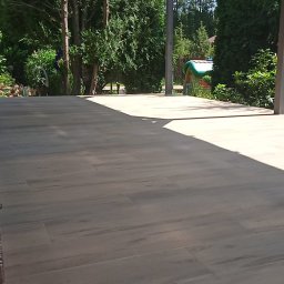 WOJT-KRAZ - Zewnętrzna drewnopodobna podłoga tarasowa pod zadaszeniem z panelami, widok na zielony ogród z drzewami iglastymi i małym domkiem dla dzieci w tle.