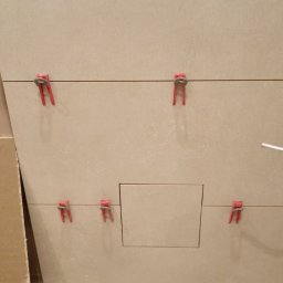 WOJT-KRAZ - Układanie beżowych płytek z plastikowymi krzyżykami dystansowymi, widoczny otwór rewizyjny, podłoga z jasnych paneli.