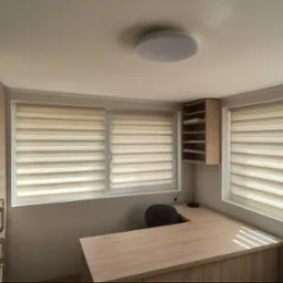 Jasne biuro z trzema oknami przysłoniętymi roletami dzień-noc, drewniane biurko narożne, szafka wisząca i wysoka zabudowa meblowa przy ścianie.