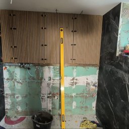 WOJT-KRAZ - Remont łazienki w trakcie: montaż paneli ściennych z drewnianych listew oraz płytek imitujących czarny marmur, widoczny poziomica i narzędzia.